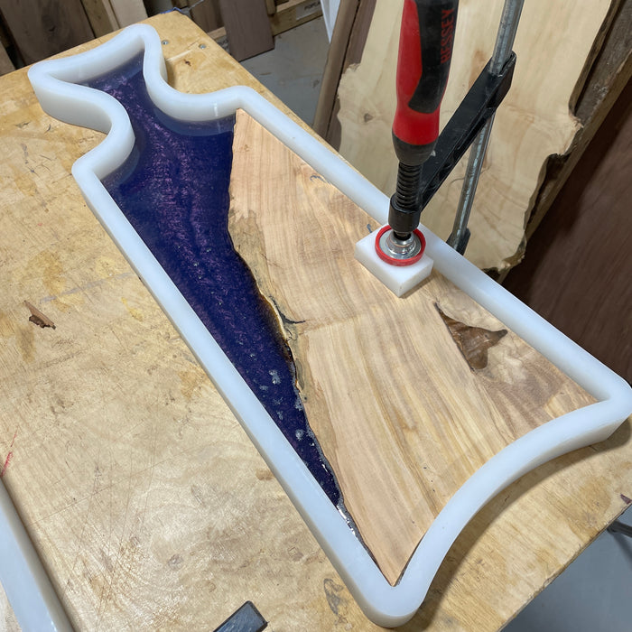 an example of an olive wood piece in a silicone mold that has a fish tale handle and purple resin poured inside. Clamp holding down the wood so it doesn't float up and and hdpe block between the clamp and wood
